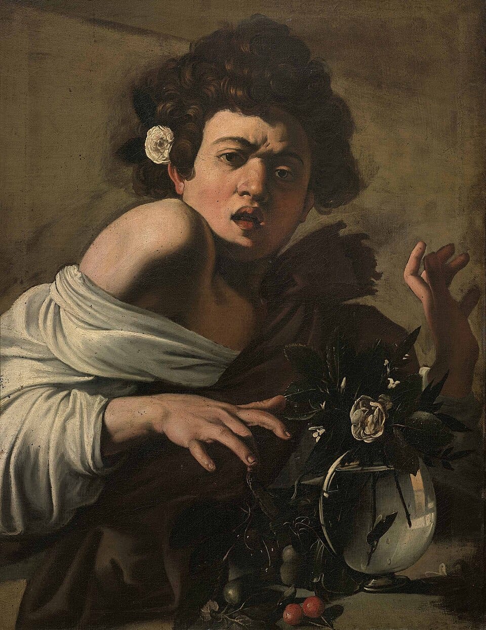 A young man with curly hair and a white rose tucked behind his ear recoils in pain, mouth open and brow furrowed, pulling his hand back from a glass vase of flowers where a lizard has bitten his finger.