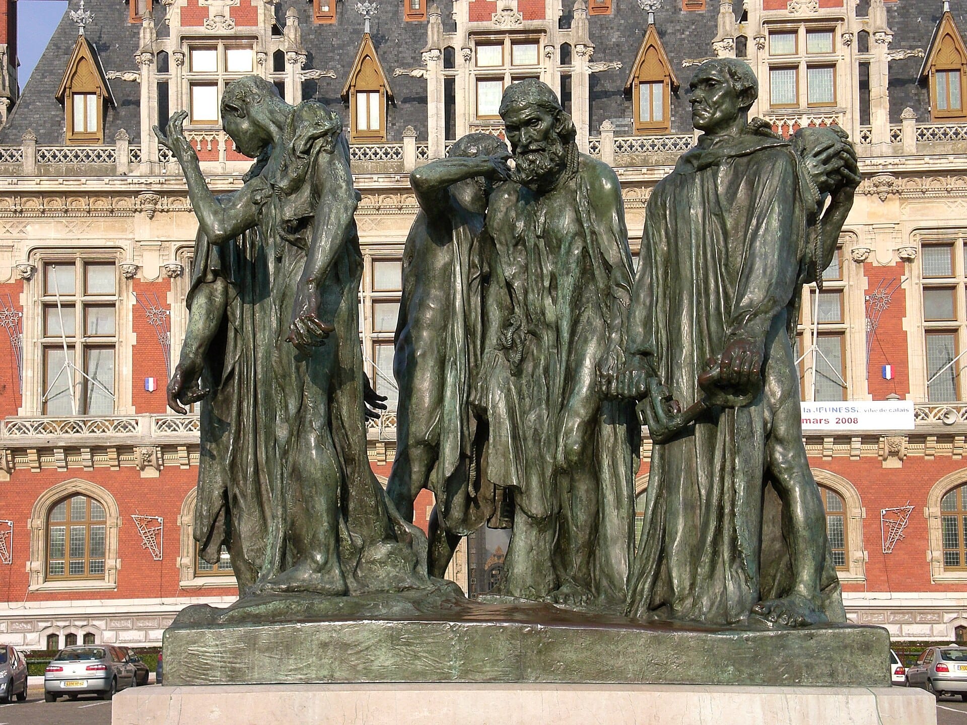 A bronze group sculpture of six middle-aged men in loose robes with ropes around their necks, standing in varied poses of resignation, grief, and resolve, displayed on a pedestal before an ornate Gothic-style municipal building.
