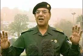 A man in a green military uniform and black beret stands outdoors, both hands raised with palms facing outward in a gesturing or emphatic pose. The background is hazy and desert-coloured. This is Mohammed Saeed al-Sahhaf, the Iraqi Information Minister known as "Baghdad Bob," photographed during a press statement in 2003.