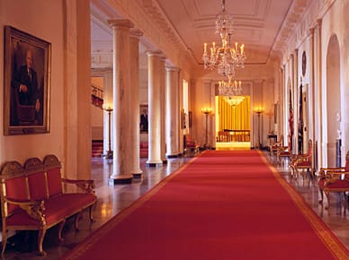 The Cross Hall of the White House, looking toward the East Room. A long red-carpeted corridor flanked by white marble columns, crystal chandeliers, and gilded chairs. A portrait hangs on the wall at the far left.