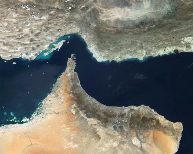 A satellite photograph of the Strait of Hormuz, the narrow waterway between Iran to the north and Oman and the United Arab Emirates to the south. The strait appears as a dark blue channel of water cutting between pale desert and rocky coastline. The Persian Gulf is visible to the left and the Gulf of Oman to the right.