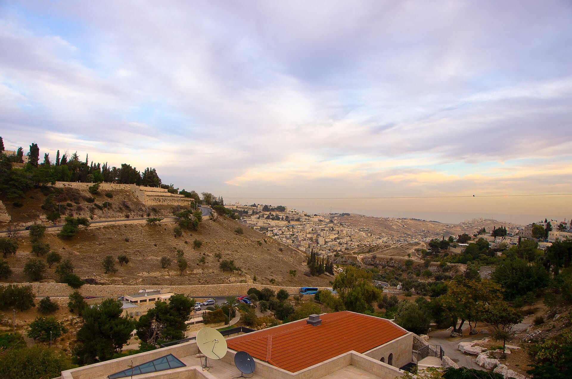 Landscape photograph of a wide terraced valley at dusk, with low stone walls along the slopes, scattered olive trees, modern white buildings on the far hillside, and a band of pink and blue sky above.