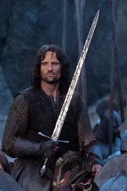 Aragorn, a long-haired warrior in dark chainmail, sits on horseback in a forest, holding a tall straight sword upright in his right hand. His expression is steady and watchful. Other mounted figures are visible behind him in soft focus.