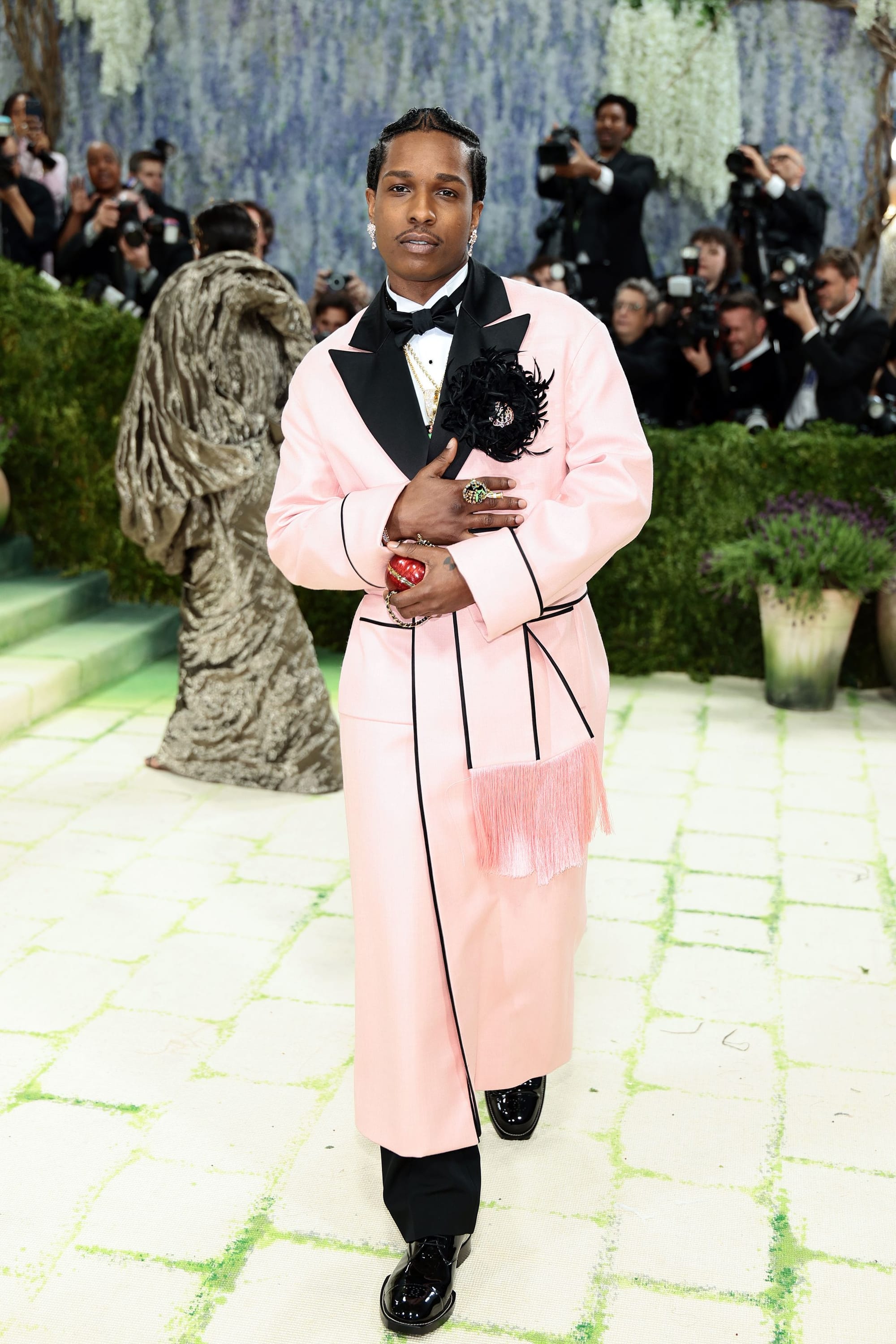 A$AP Rocky at the 2026 Met Gala, photographed on the red carpet in a long pink wool coat with black satin lapels and piping, accented by a black silk-and-feather camellia brooch, worn over a white shirt, black bow tie, and black trousers with patent leather shoes. Photographers and a backdrop of cascading white wisteria visible behind him.