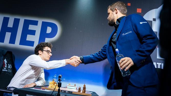 Two players shake hands across a chess table at a tournament, with a sponsor backdrop visible behind them.
