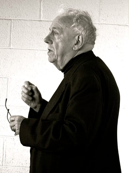 Black-and-white photograph of Alasdair MacIntyre in profile, an elderly man with disheveled white hair, wearing a dark jacket, mid-speech with one hand raised in gesture and the other holding his glasses, against a plain cinderblock wall.