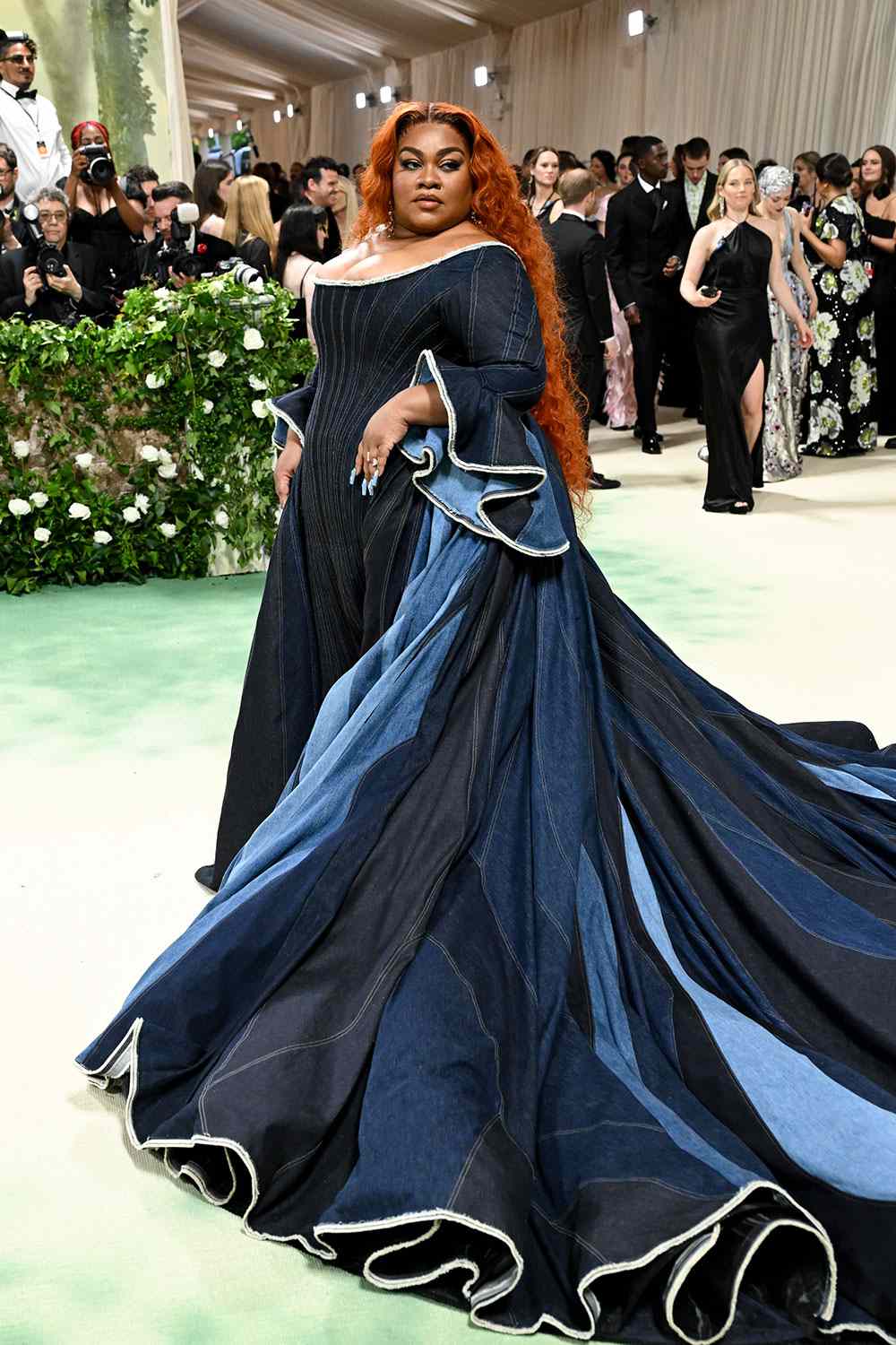 Da'Vine Joy Randolph at the 2024 Met Gala in a structured corseted ball gown made entirely of patchwork denim in varying shades of indigo, with an off-shoulder neckline, scalloped white-trimmed sleeves, and a sweeping flowing train. Her long copper-red hair falls past her shoulders. Photographers and other guests visible in the background.