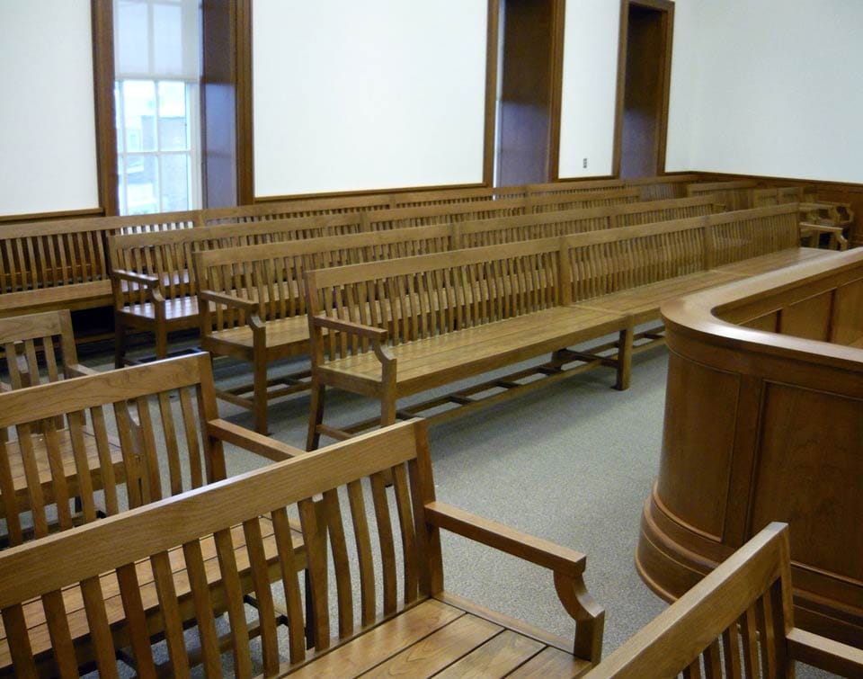 An empty courtroom interior showing wooden gallery benches in two rows, a wooden jury box railing at right, and tall windows; the room is unoccupied.