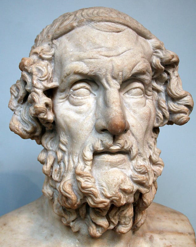 A marble bust of an elderly bearded man with curly hair and a thick curled beard, his sightless eyes turned slightly upward. The surface of the stone is weathered, with some chipping at the nose and ear. The bust is photographed against a plain light-blue background.