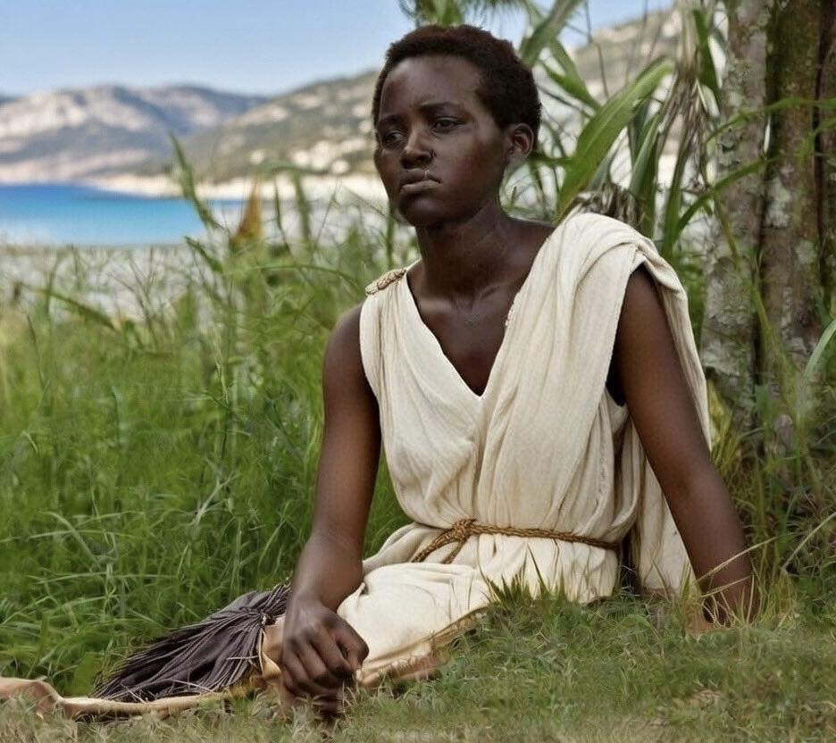 A young woman with close-cropped dark hair sits on grass beneath a tree, leaning lightly against the trunk. She wears a simple unbleached sleeveless chiton belted at the waist with a rope cord. Behind her is a vivid blue sea and pale hills under a clear sky.