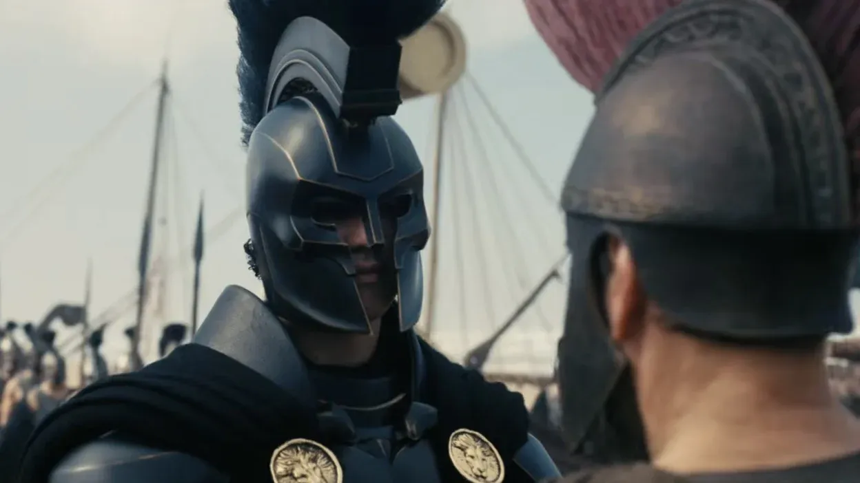 A young warrior in a black muscle-cuirass faces an older bearded warrior in profile. He wears a dark Corinthian-style helmet with a tall black horsehair crest, and his cuirass bears two large gold medallions embossed with lion heads. The masts and rigging of beached ships are visible behind him.