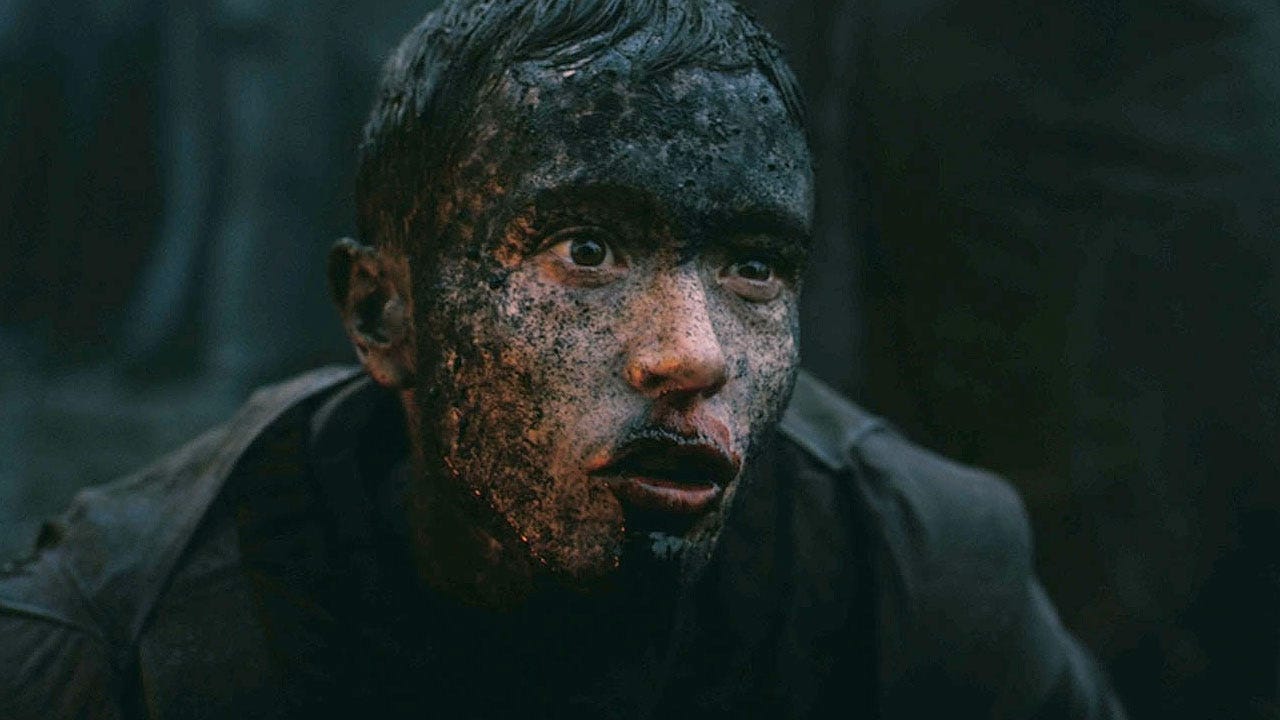 A close-up of a young person with short dark hair, their face heavily streaked with grey mud and dirt. Their eyes are wide and fixed on something out of frame. The lighting is dim and bluish, suggesting a cave or underworld setting.