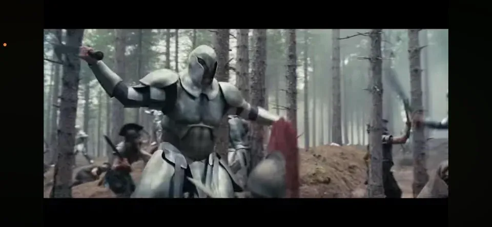 A massive armored figure looms in a misty pine forest, dwarfing the smaller human warriors fighting around its legs. The figure wears full articulated plate armor — segmented shoulder, chest, and thigh pieces — and a tall fin-crested helmet that covers the face. Its arms are raised, wielding a weapon overhead. Other warriors with spears and red-plumed helmets are visible in the background.