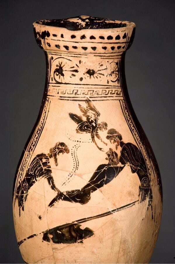 A bulbous ancient Greek ceramic vase with a flared mouth, decorated in black-figure on the pale clay ground. Bands of ornament run around it: a row of dots at the lip, a floral rosette on the shoulder, and a meander (Greek key) band above the main panel. In the scene, two dark silhouetted figures crouch facing each other over a reclining figure between them, while a small winged figure hovers in the upper field above them. The surface is cracked and worn, and the vase is mounted against a plain grey background.