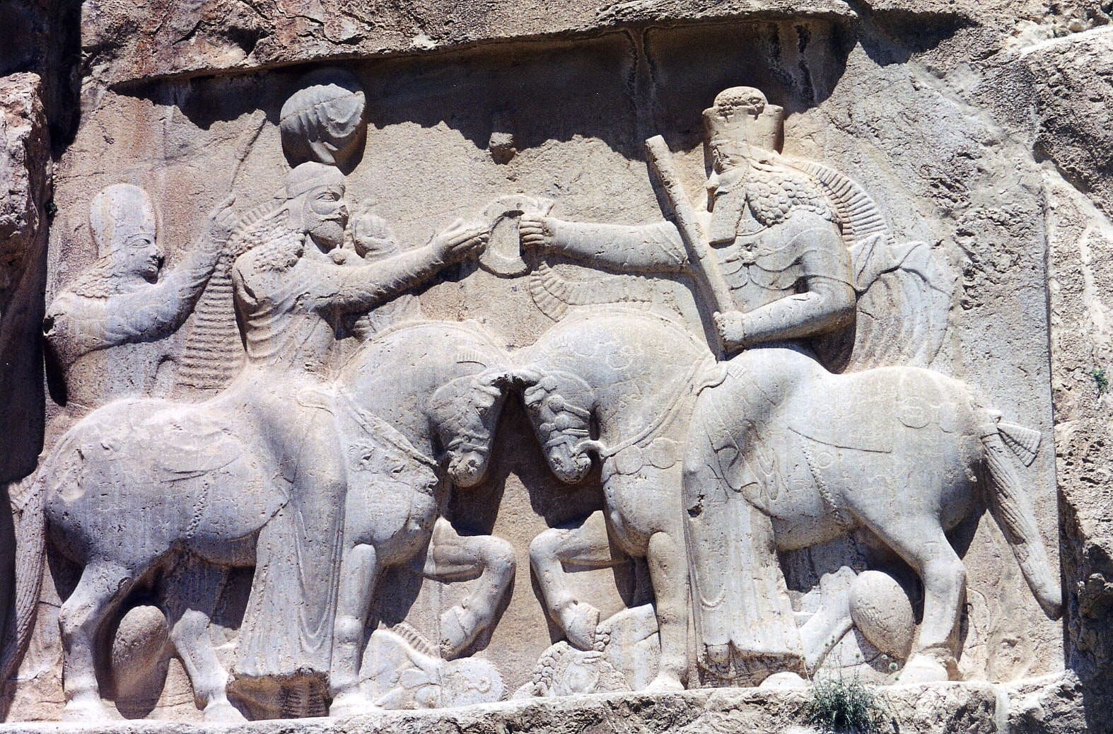 A weathered rock relief of two crowned figures on horseback facing each other, the right-hand figure passing a ring to the left, defeated enemies crushed beneath both horses.