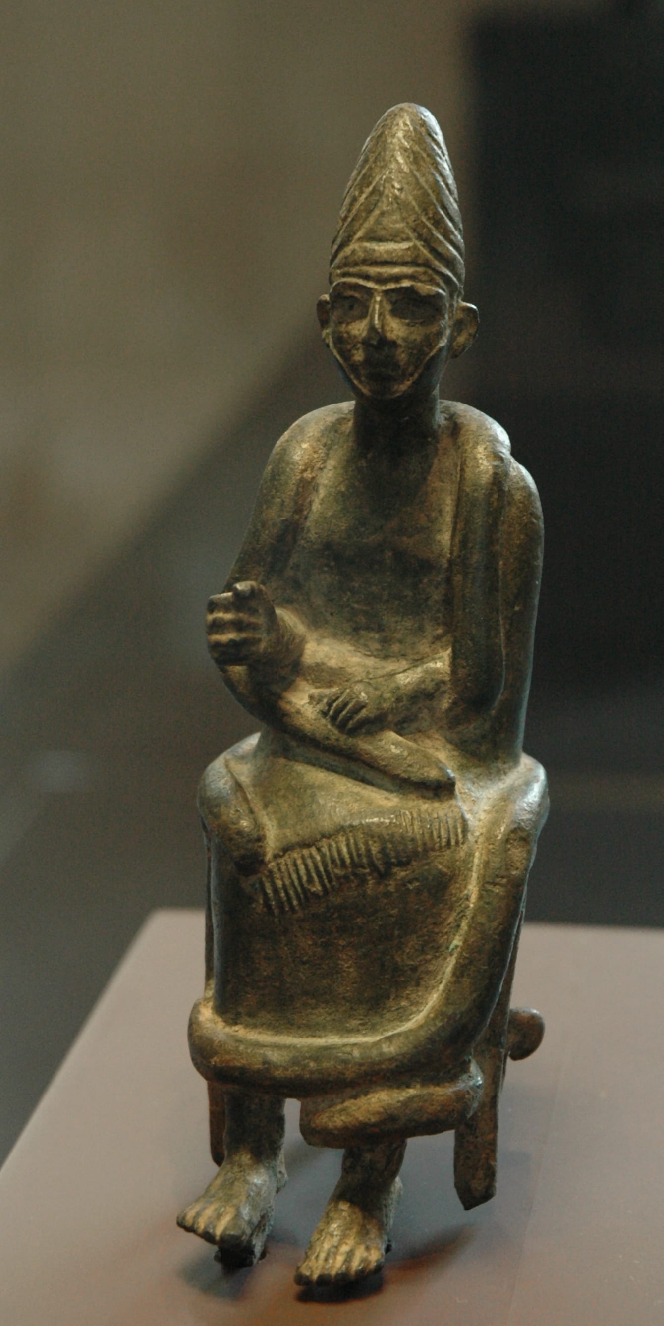  Small Late Bronze Age bronze figurine of a seated bearded deity with a tall conical crown, one hand raised in blessing, identified as the Canaanite high god El from Ugarit.