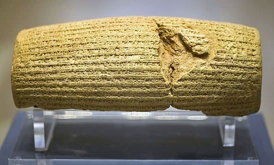 A barrel-shaped clay cylinder covered in rows of Akkadian cuneiform, cracked across the middle, displayed on a clear acrylic stand in a museum case.