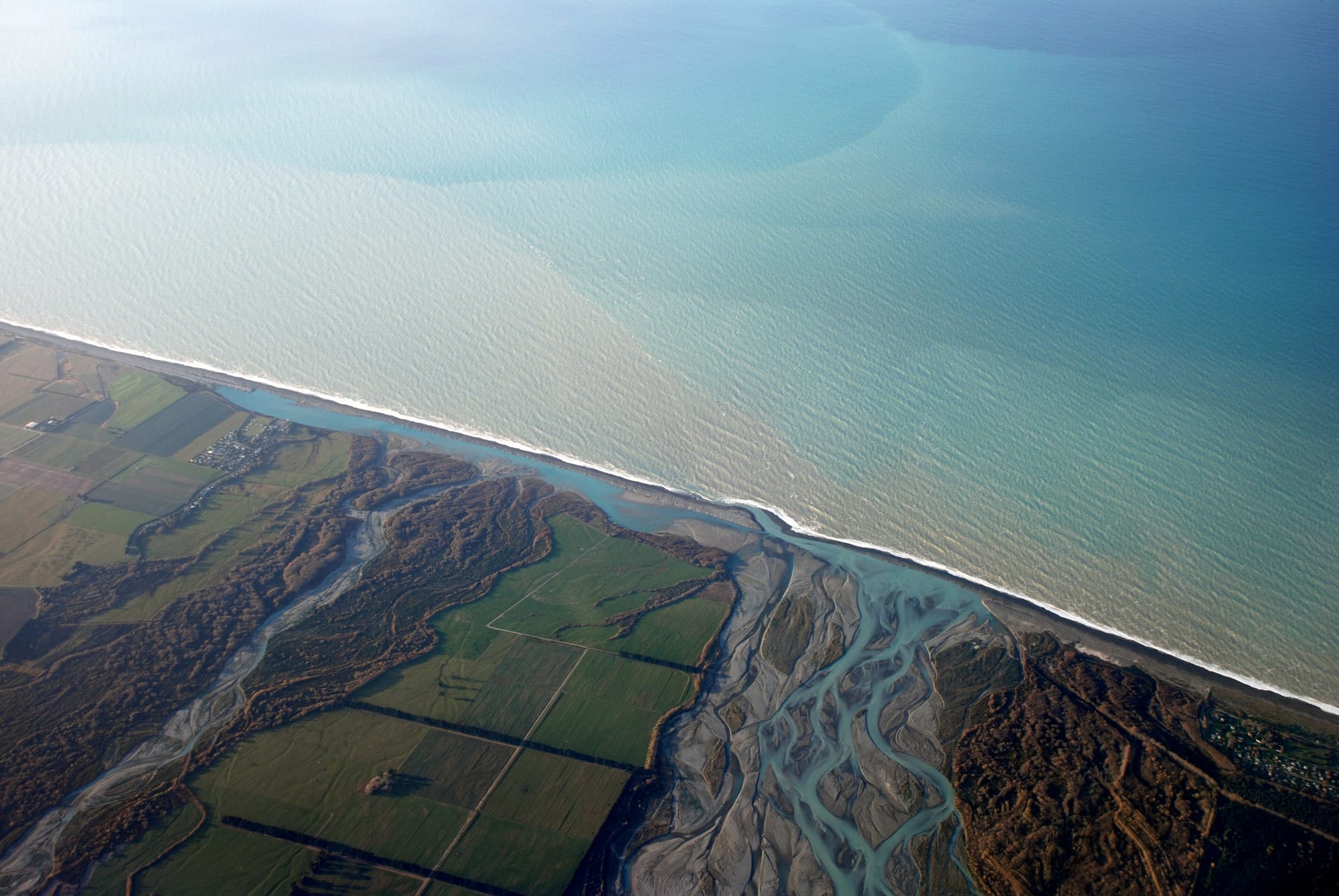 Aerial photograph of a braided river's channels crossing a gravel plain and merging into the ocean, sediment plume visible in the sea.