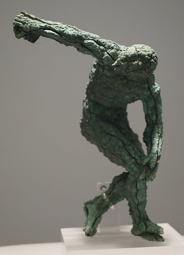 A fragmentary ancient Roman bronze statue of a discus thrower in mid-throw pose, its surface heavily cracked and patinated green.