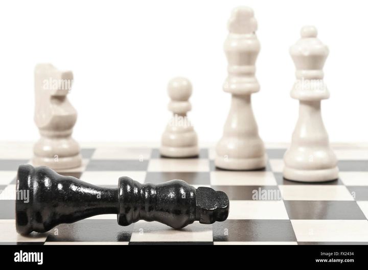 A black king lies on its side on a chessboard while four white pieces stand upright in soft focus behind it.