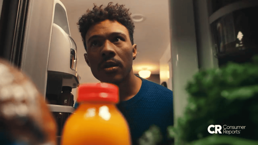 A young man peers into an open refrigerator with a puzzled expression, shot from inside the fridge looking out, with a bottle of orange juice in the foreground. Consumer Reports logo in the bottom right corner.