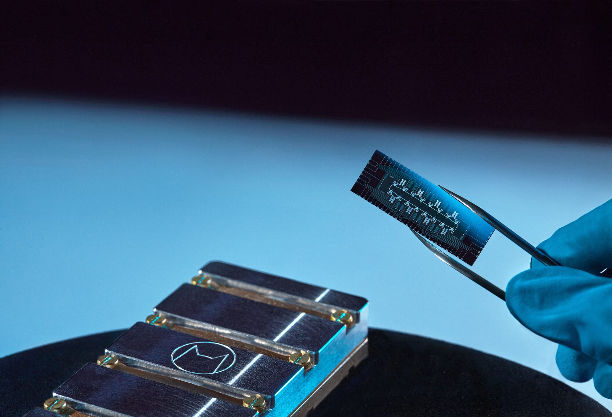 Une puce supraconductrice à qubits-chats d'Alice & Bob tenue à la pince, au-dessus de son boîtier métallique frappé du logo