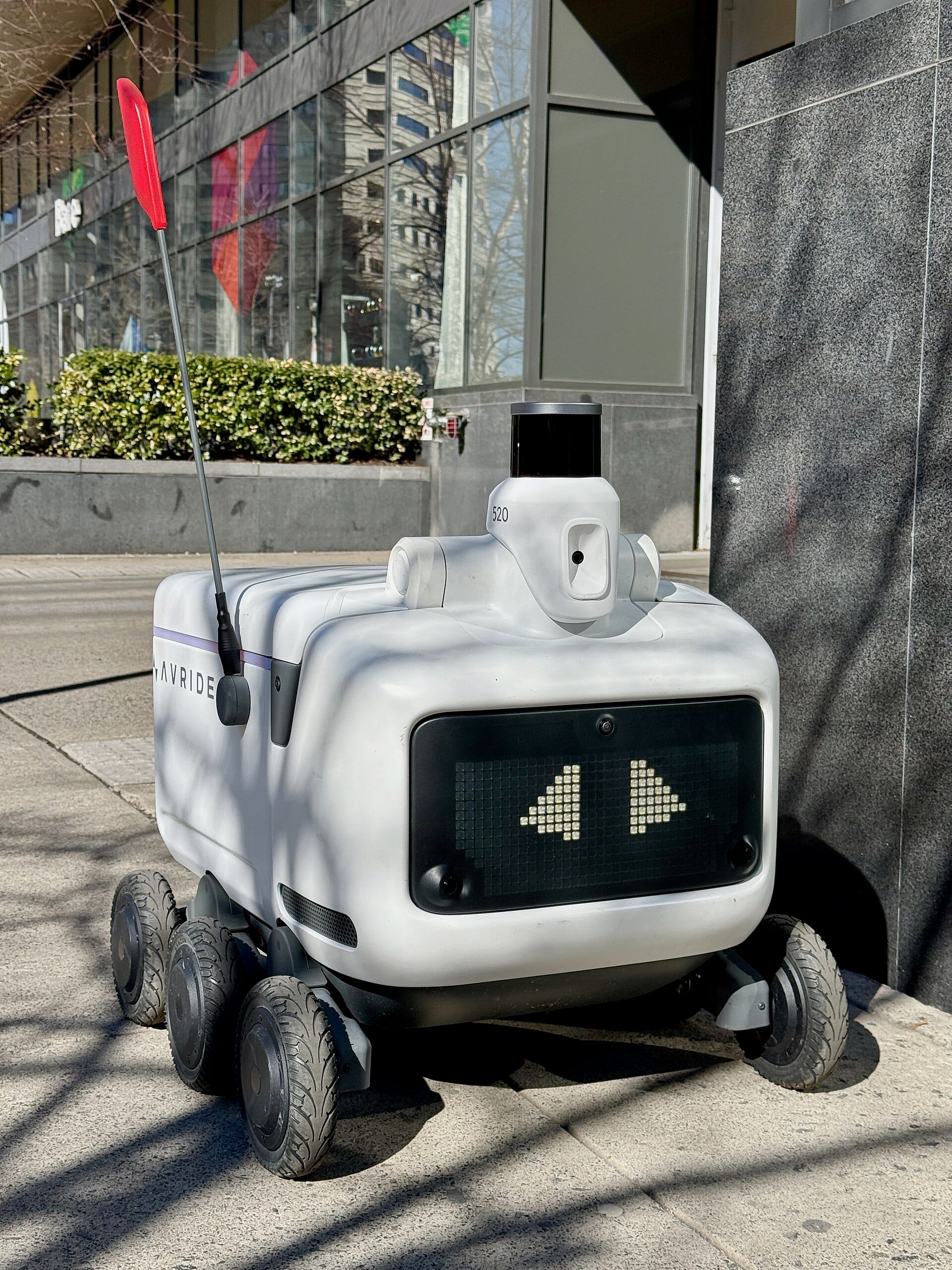 Un robot de livraison autonome Avride, avec son drapeau rouge, son lidar et ses yeux lumineux, sur un trottoir au pied d'un immeuble vitré