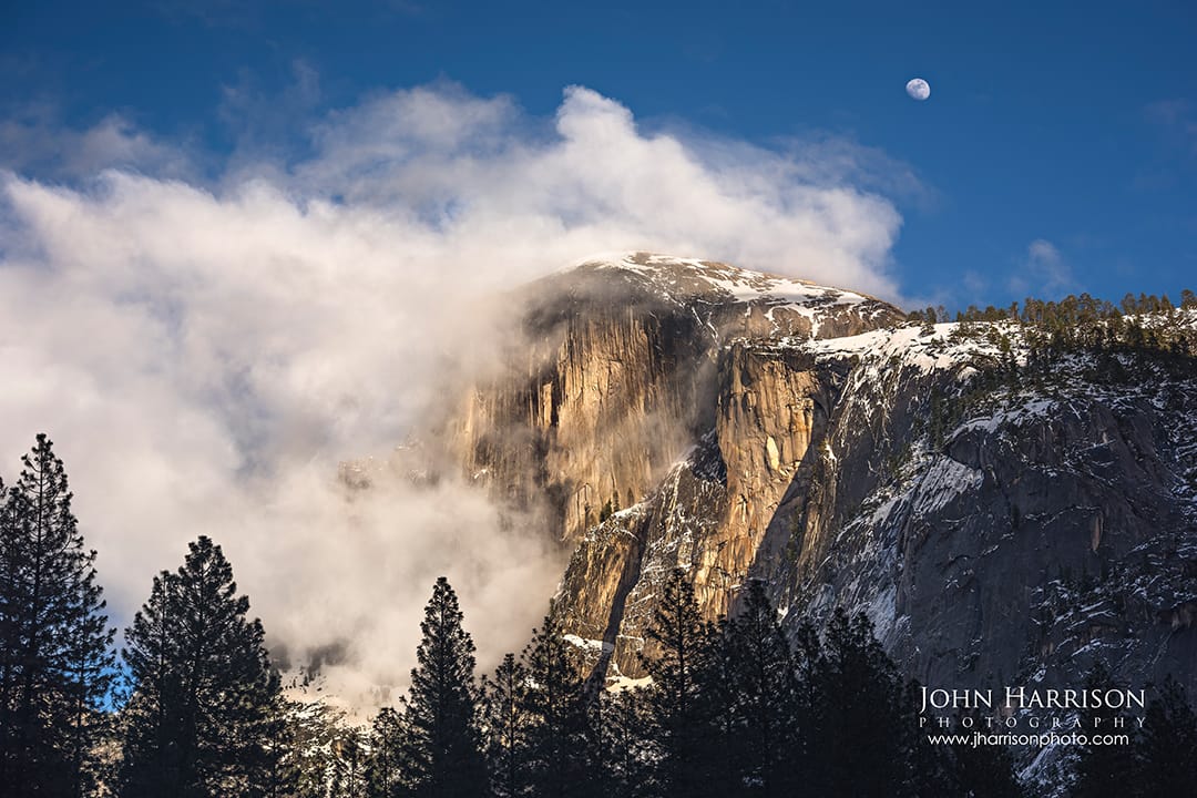 Half Dome emerging through winter clouds with snow-dusted granite and a rising moon above Yosemite National Park, California