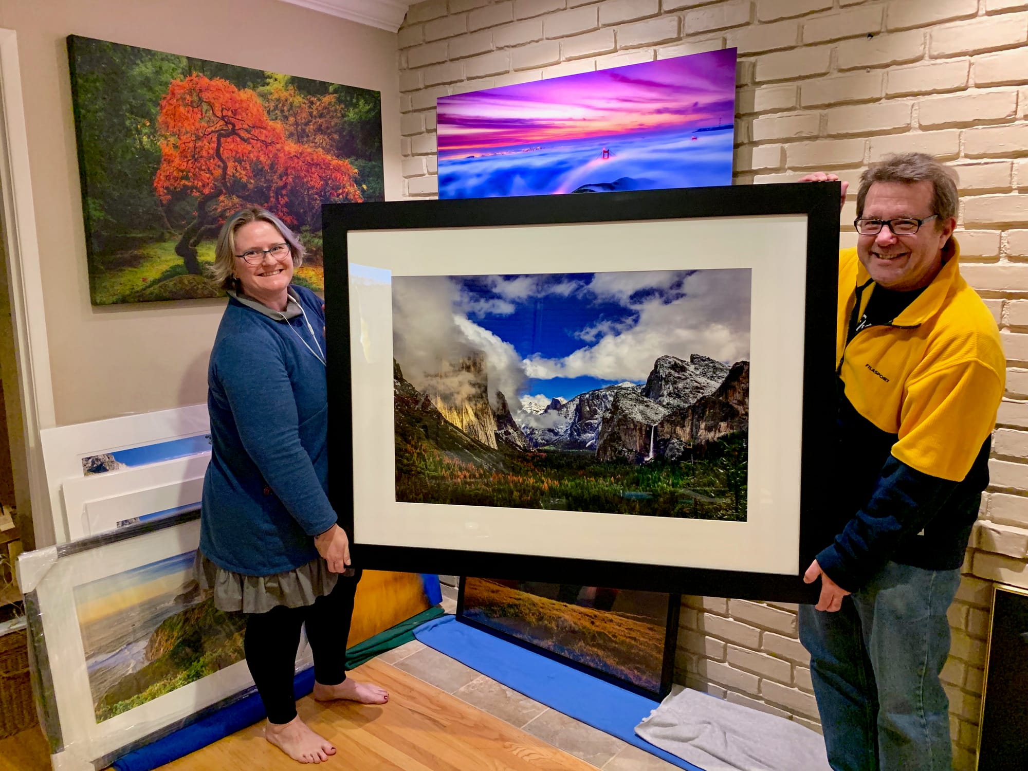 A smiling couple holding a large black-framed fine art print of Yosemite’s Tunnel View in winter, displayed in their home alongside additional landscape photography