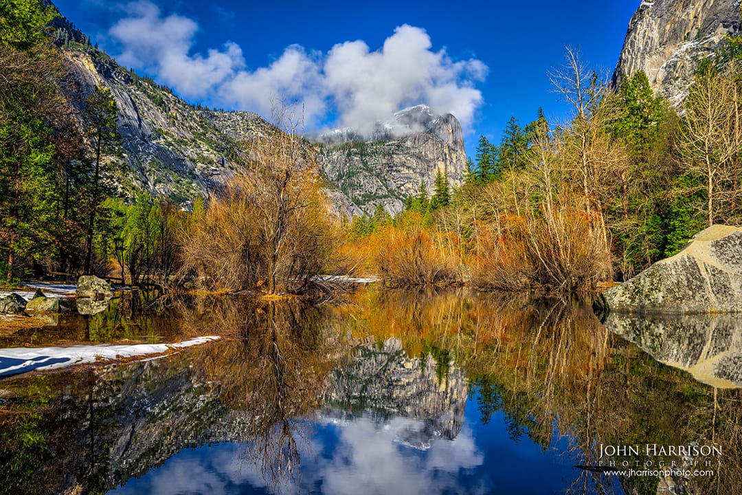 Reflection of Mount Watkins and Yosemite Valley granite cliffs in calm water at Mirror Lake, with autumn color trees, blue sky, and drifting clouds in Yosemite National Park, California.