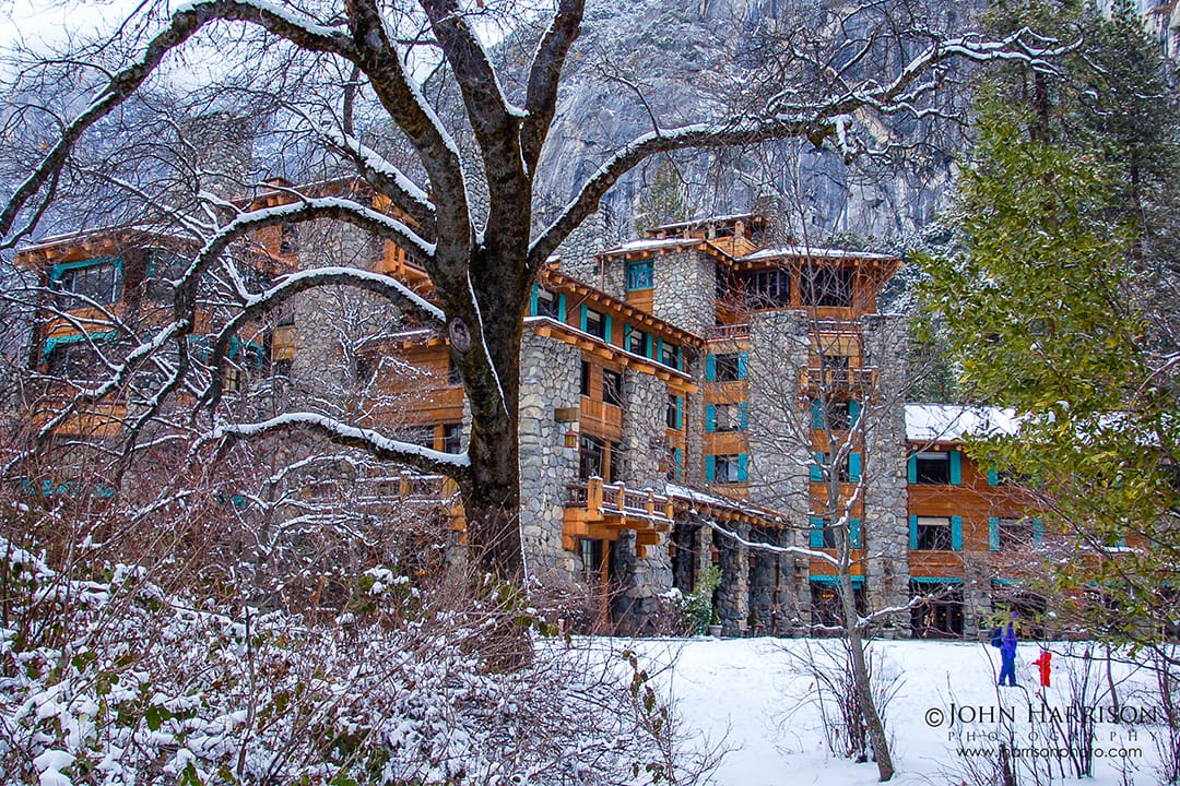 The Ahwahnee Hotel in Yosemite Valley covered in fresh snow, framed by winter trees with granite cliffs rising behind in Yosemite National Park, California.