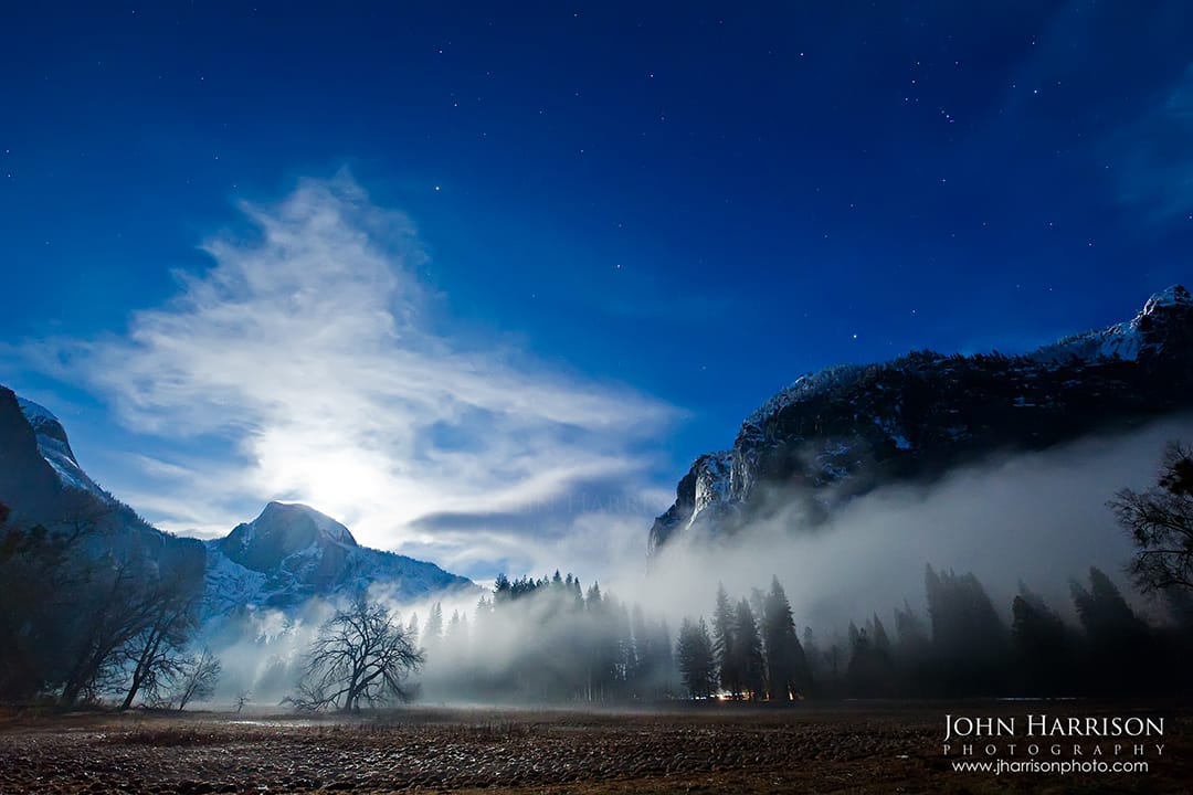 Full moon rising behind Half Dome with blue night sky, swirling fog and mist across Yosemite Valley meadow, and moonlit clouds over Yosemite National Park, California.