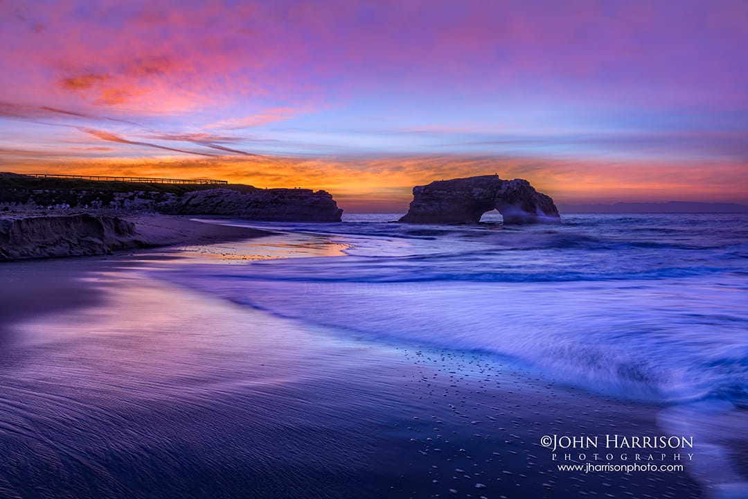 Colorful sunrise at Natural Bridges State Beach in Santa Cruz, California with long exposure waves, wet sand reflections, and the Natural Bridges rock arch in the Pacific Ocean.