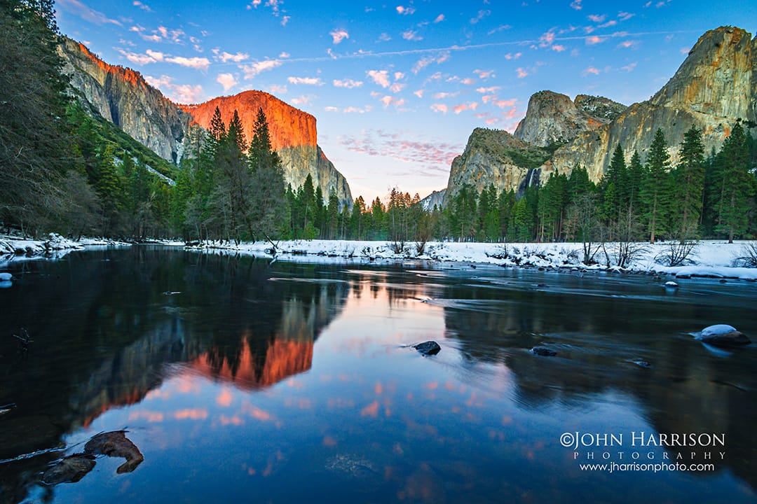Winter sunset at Valley View in Yosemite National Park with El Capitan glowing orange above the Merced River, snow-covered riverbanks, and reflections in Yosemite Valley.