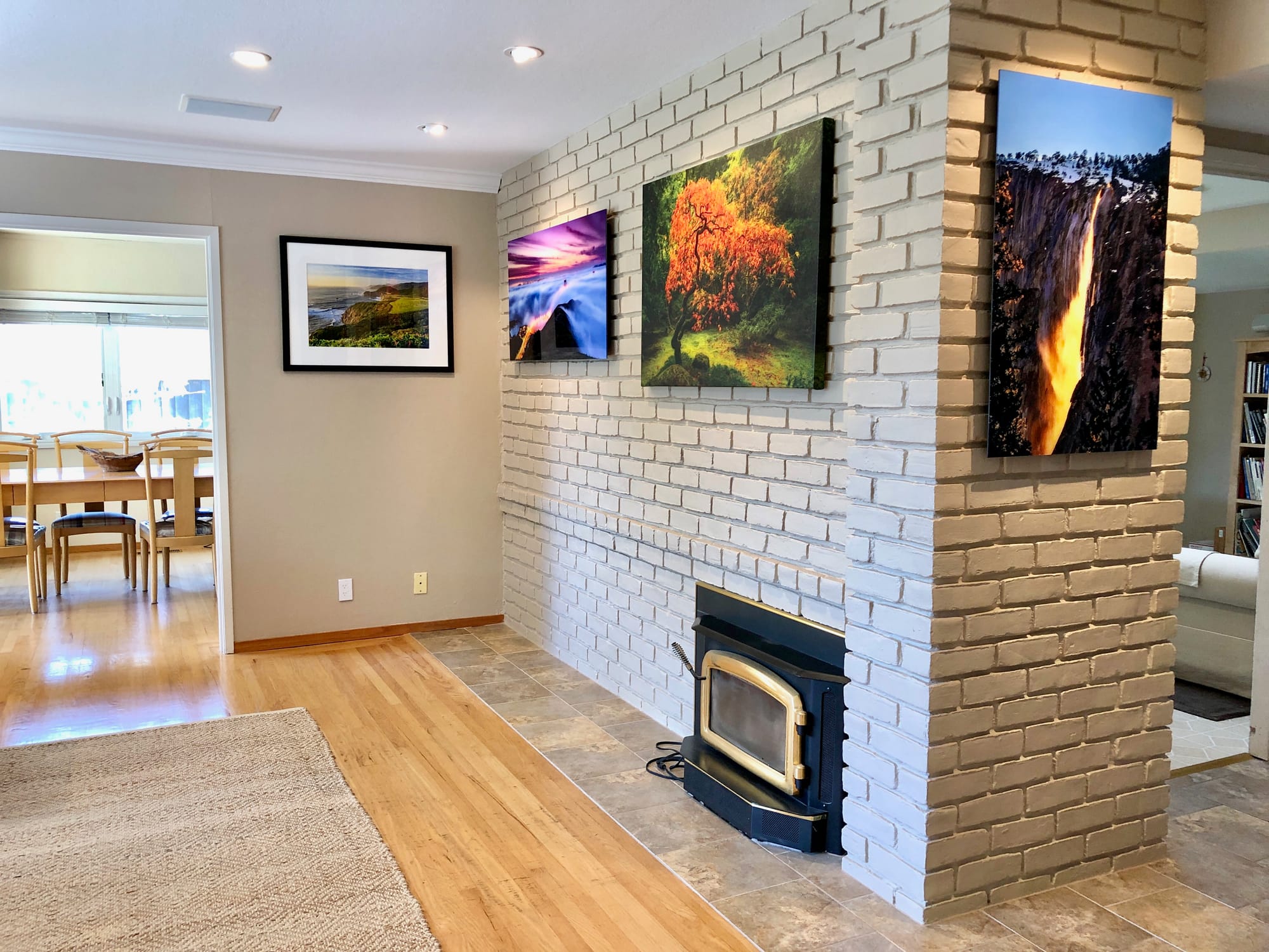 Fine art photography installation featuring Japanese maple, coastal seascape, and Yosemite Firefall prints displayed on a white brick fireplace wall in a modern residential interior.
