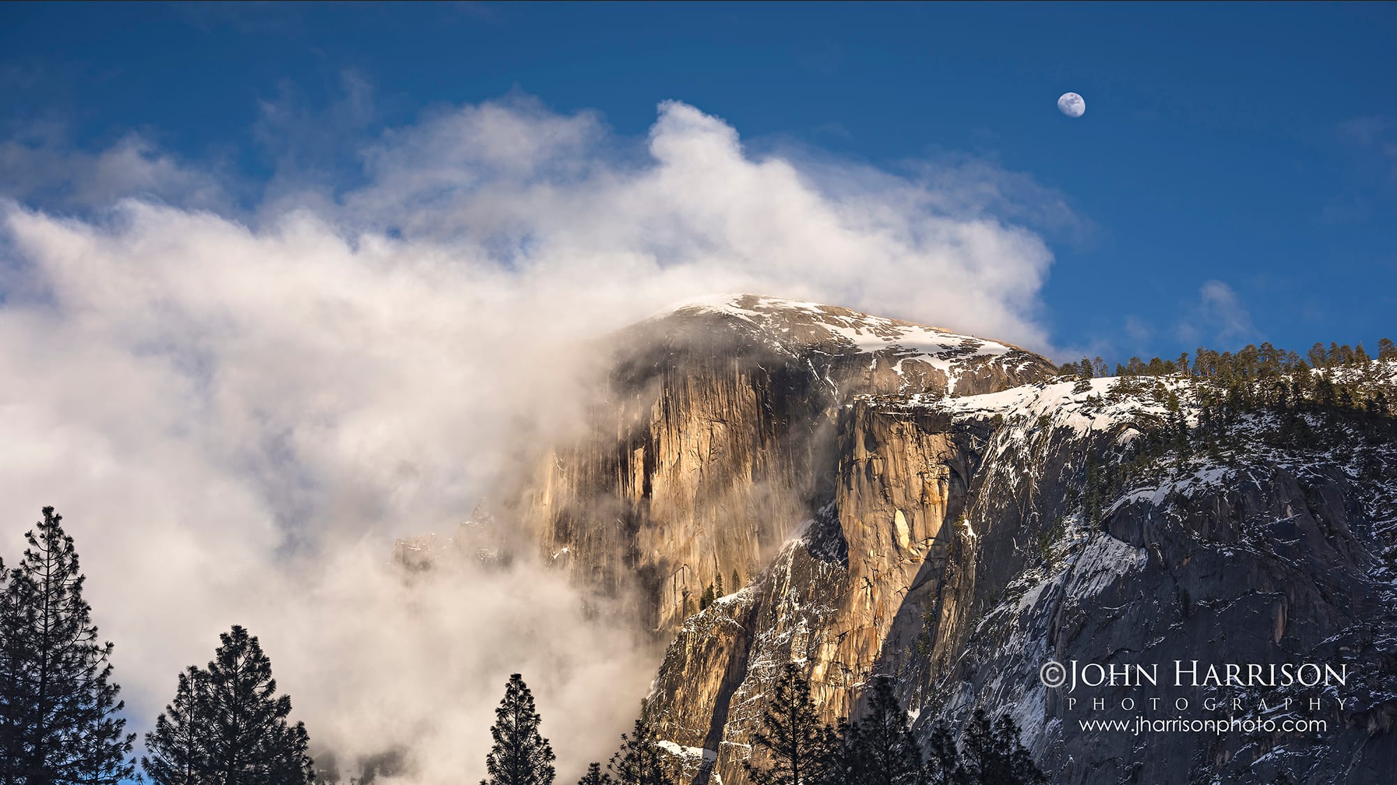 Half Dome in Yosemite National Park partially illuminated by winter sunlight as storm clouds and mist wrap around the granite dome, with snow on the summit and pine trees silhouetted in the foreground.