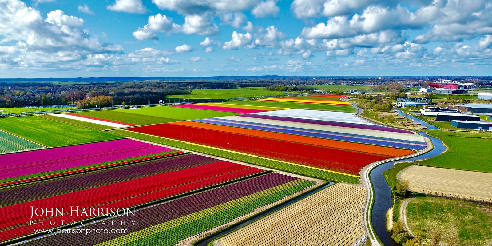 Aerial photography of colorful tulip fields and winding canals near Alkmaar, North Holland. Symmetrical rows of red and purple flowers seen from above in the Dutch polders during spring 2026.
