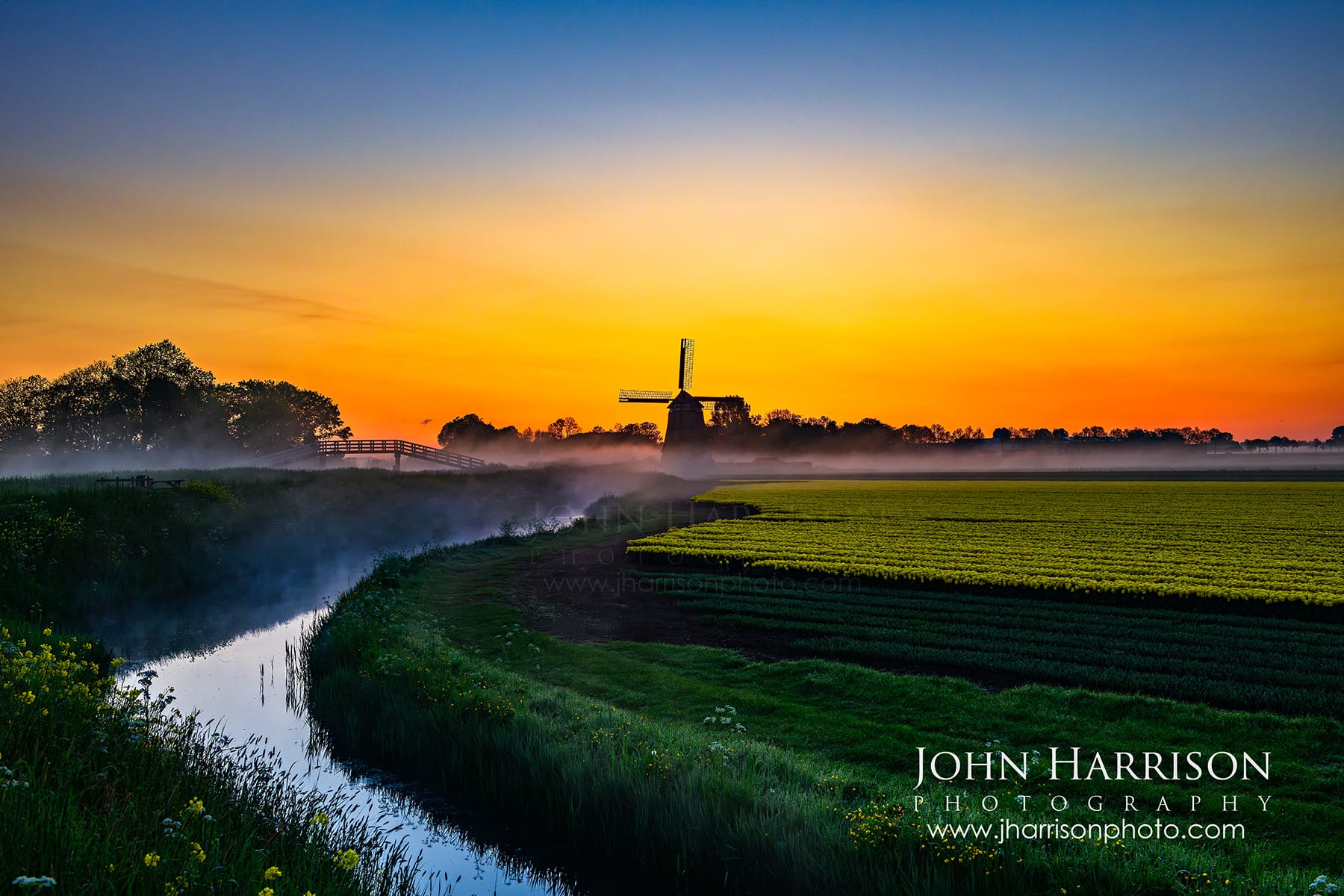 Sunrise at Obdammermolen windmill near Obdam. A 17th-century thatched polder mill silhouetted against an orange sky with morning mist over green tulip fields and a canal in North Holland.