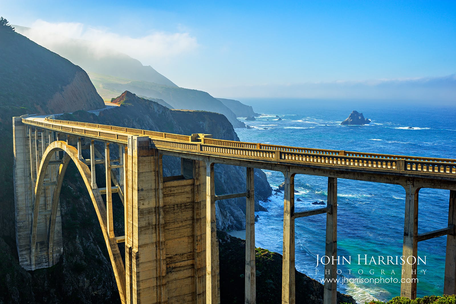Bixby Bridge spanning Bixby Creek Canyon in Big Sur California with turquoise Pacific Ocean water, coastal cliffs, and marine fog in the distance.