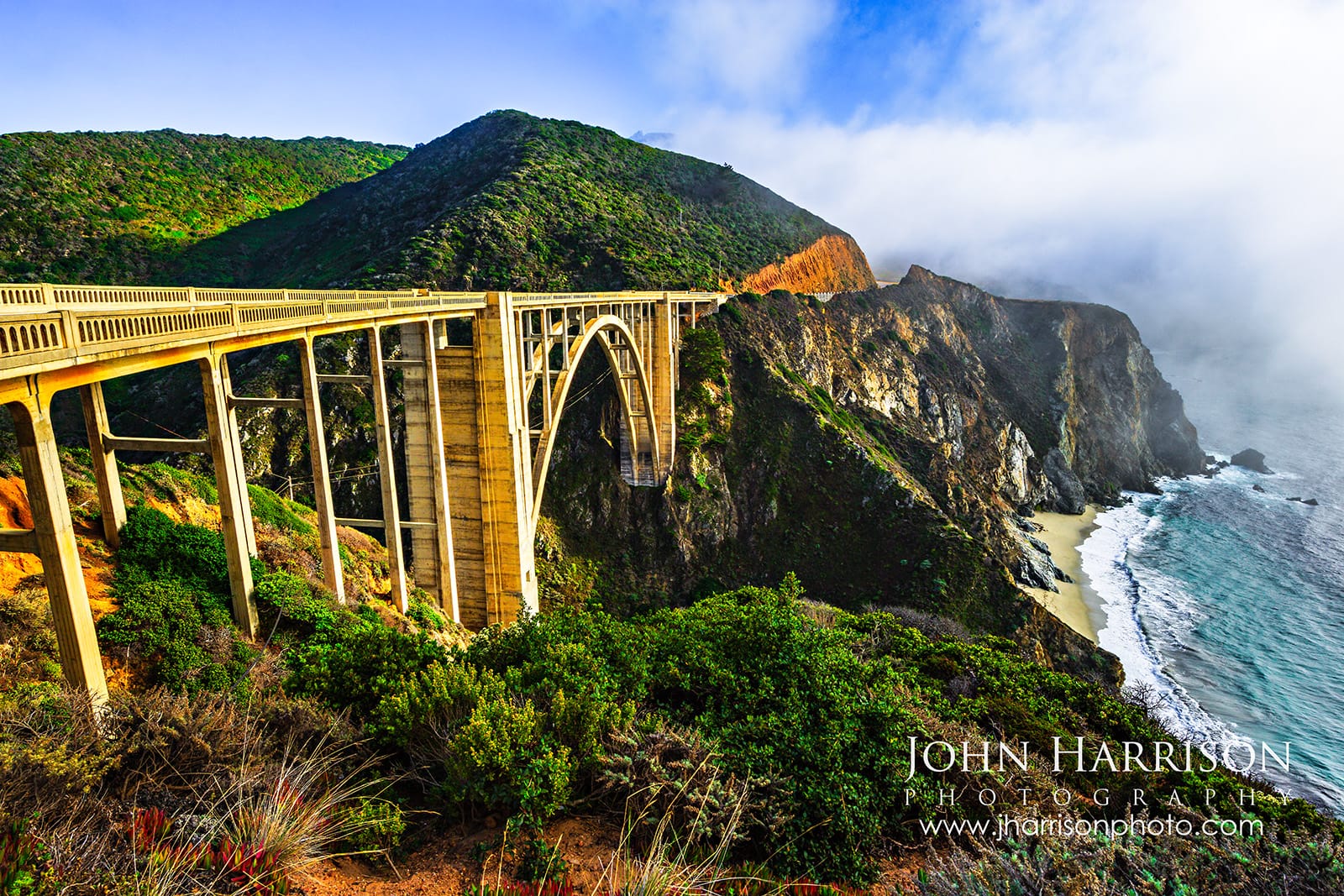 Bixby Bridge in Big Sur California illuminated by late afternoon sunlight with coastal fog, green hillsides, rugged cliffs, and the Pacific Ocean below.