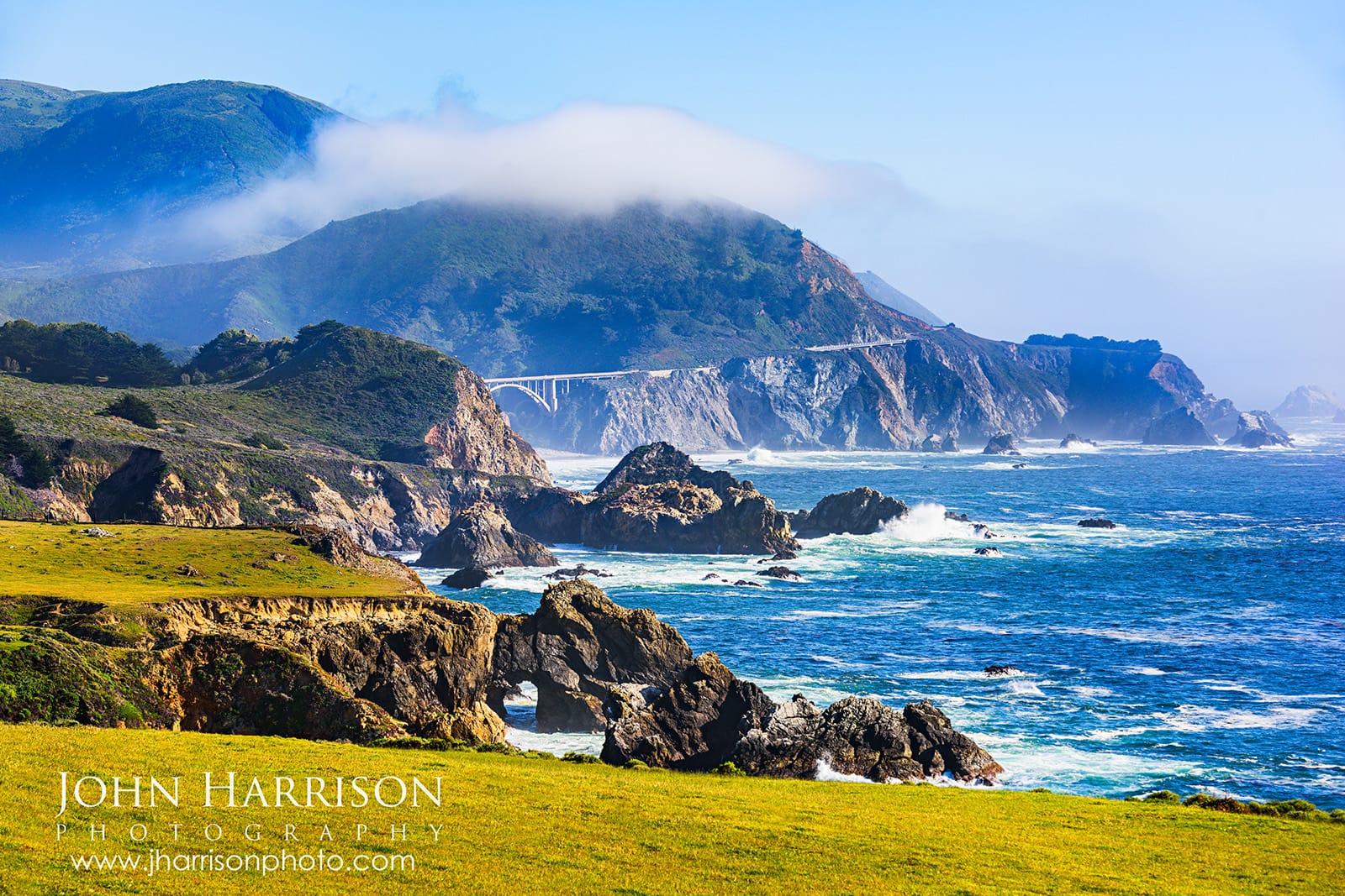 Natural rock arch along the Big Sur coastline near Garrapata State Park with Bixby Bridge in the distance, coastal fog rolling over the hills, and Pacific Ocean waves below.