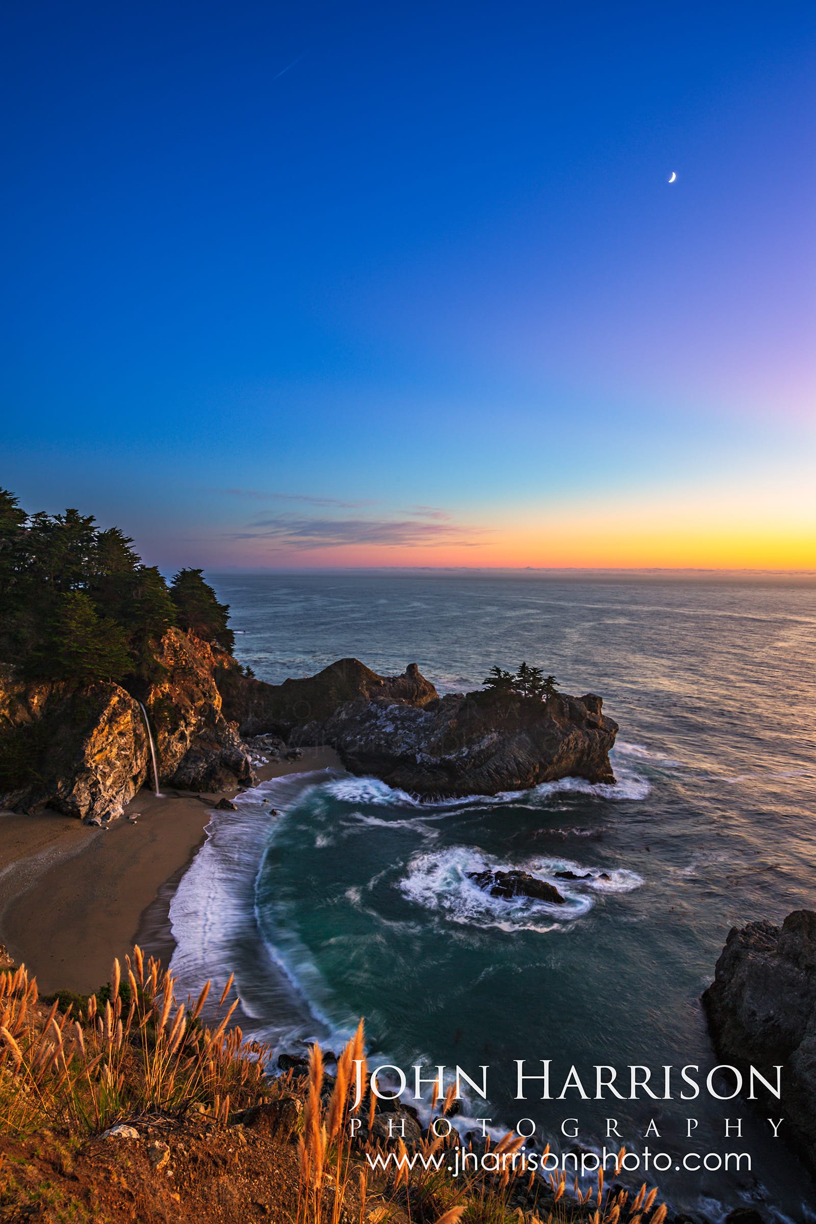 Crescent moon rising above McWay Falls at twilight in Julia Pfeiffer Burns State Park, Big Sur, California, with the Pacific Ocean and rugged Highway 1 coastline illuminated by fading evening light.
