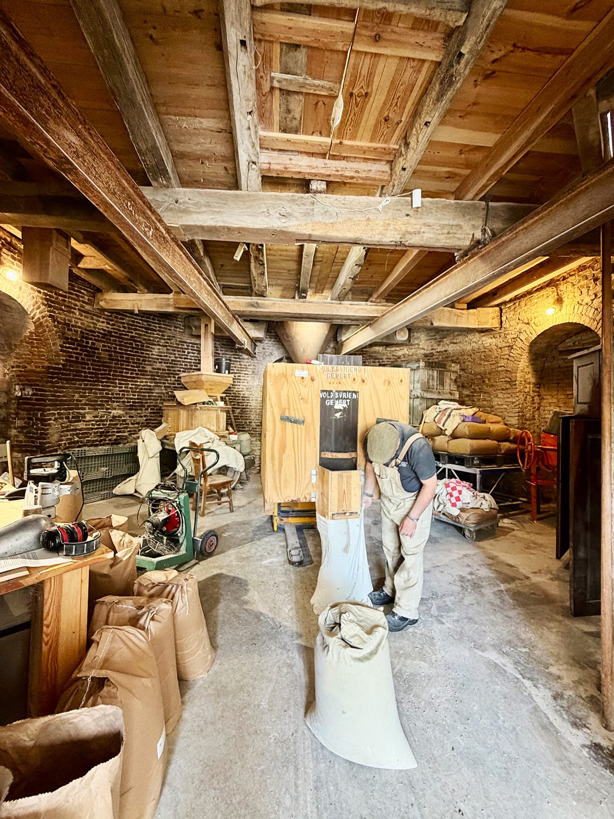 Miller filling sacks with freshly ground flour inside Molen De Volksvriend windmill in Gemert near Eindhoven, surrounded by historic wooden beams, brick walls, and traditional milling equipment.