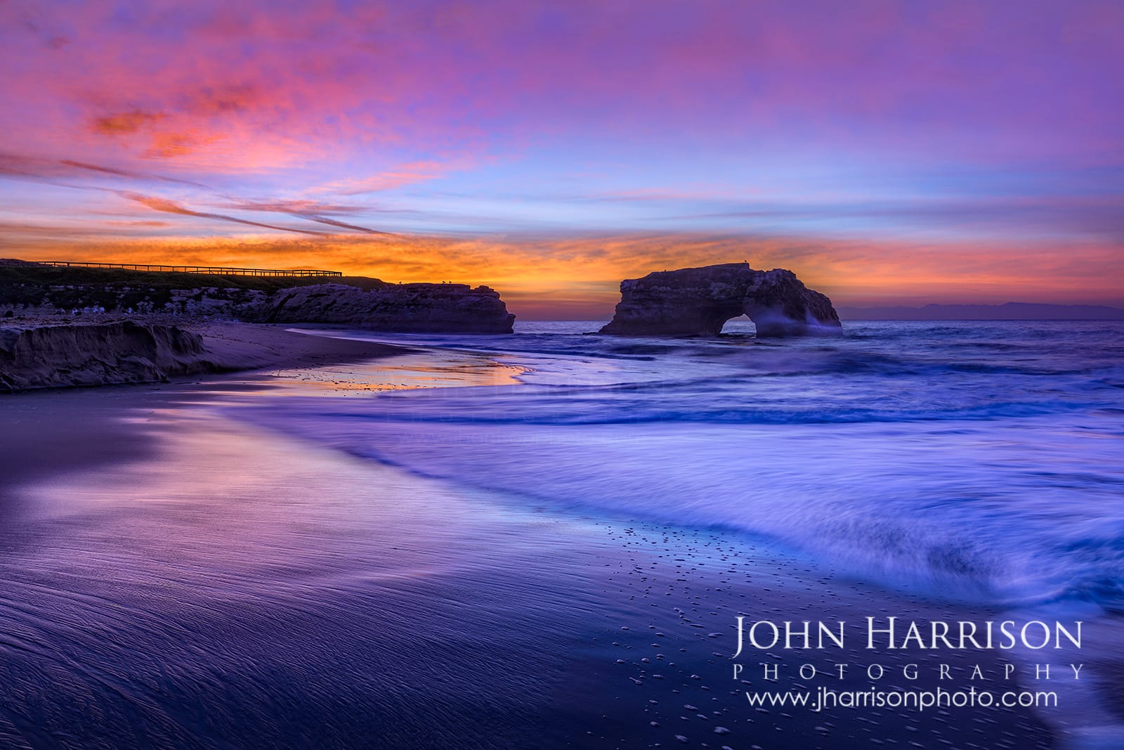 Colorful sunrise at Natural Bridges State Beach in Santa Cruz, California with long exposure waves, wet sand reflections, and the Natural Bridges rock arch in the Pacific Ocean.