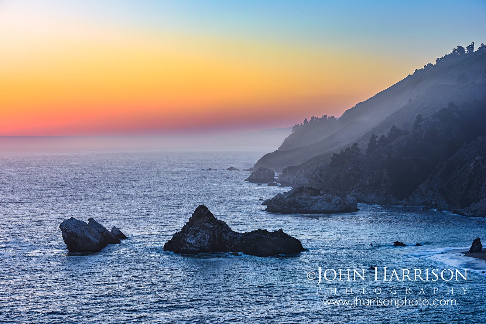 Sunset glow over the Northern California coastline near Big Sur with coastal seastacks, layered cliffs, ocean haze, and pastel evening skies along Highway 1.