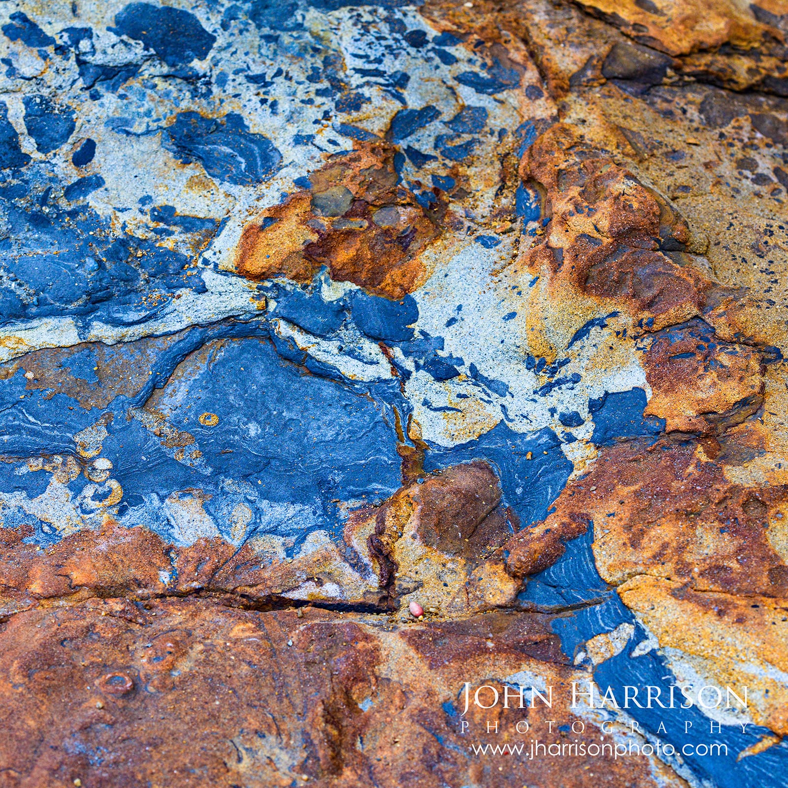 Painted by Nature, a square fine art photograph by John Harrison revealing natural blue-gray, rust, gold and ochre rock textures at Weston Beach in Point Lobos State Natural Reserve, California.