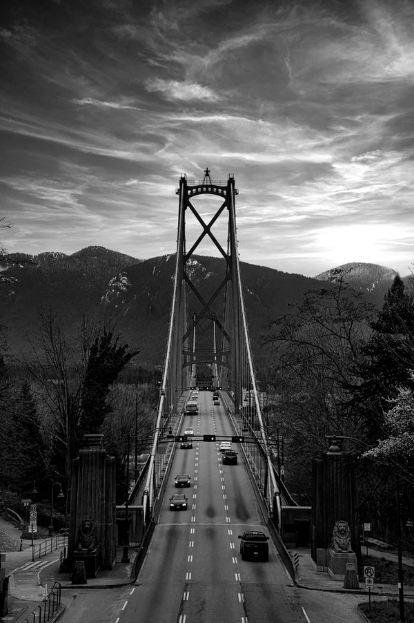 View looking down the Lions Gate Bridge roadway in Vancouver, with lion sculptures at the entrance, suspension cables overhead, cars in traffic, and forested mountains in the background