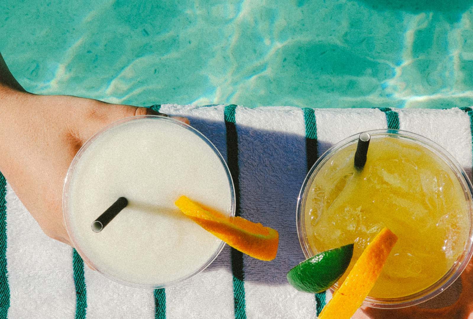 Two hands holding tropical drinks with orange and lime garnishes on a striped pool towel beside a clear pool, capturing a relaxing summer moment after ensuring pool safety.