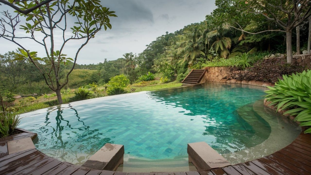 An infinity-edge eco-friendly pool with turquoise water, surrounded by wooden decking and lush green hills in the background.