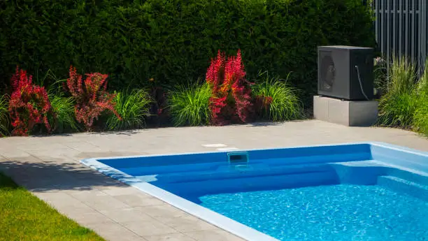 A compact pool heat pump unit installed near a residential pool, surrounded by greenery and flowers in a sunny backyard.